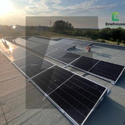 Instalacja paneli słonecznych na płaskim dachu z widocznym zachodzącym słońcem. Panele ułożone w rzędach, z widocznymi elementami montażowymi i krajobrazem w tle.