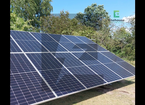 Panel słoneczny na tle zieleni i błękitnego nieba. Widoczne moduły fotowoltaiczne z logo firmy Enehouse w prawym górnym rogu. Instalacja na trawniku.