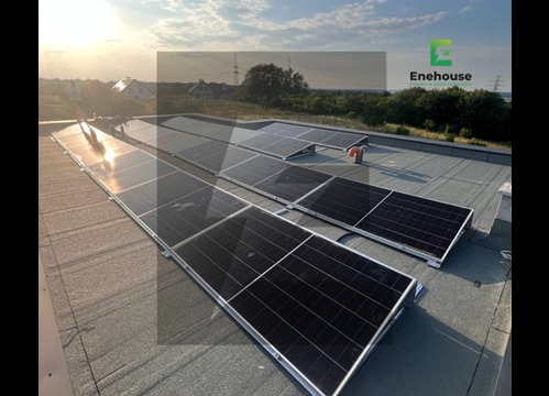 Instalacja paneli słonecznych na płaskim dachu z widocznym zachodzącym słońcem. Panele ułożone w rzędach, z widocznymi elementami montażowymi i krajobrazem w tle.