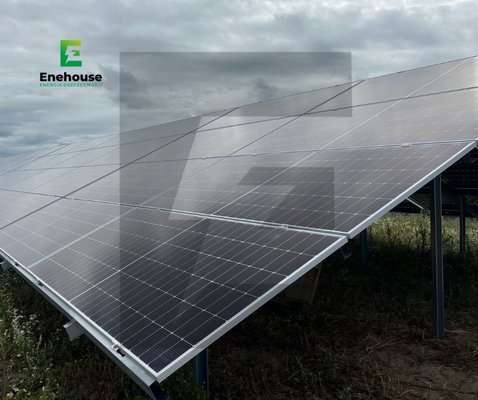 Panel słoneczny pod pochmurnym niebem, widoczny z dołu pod kątem. Logo firmy w lewym górnym rogu. Instalacja na łące.