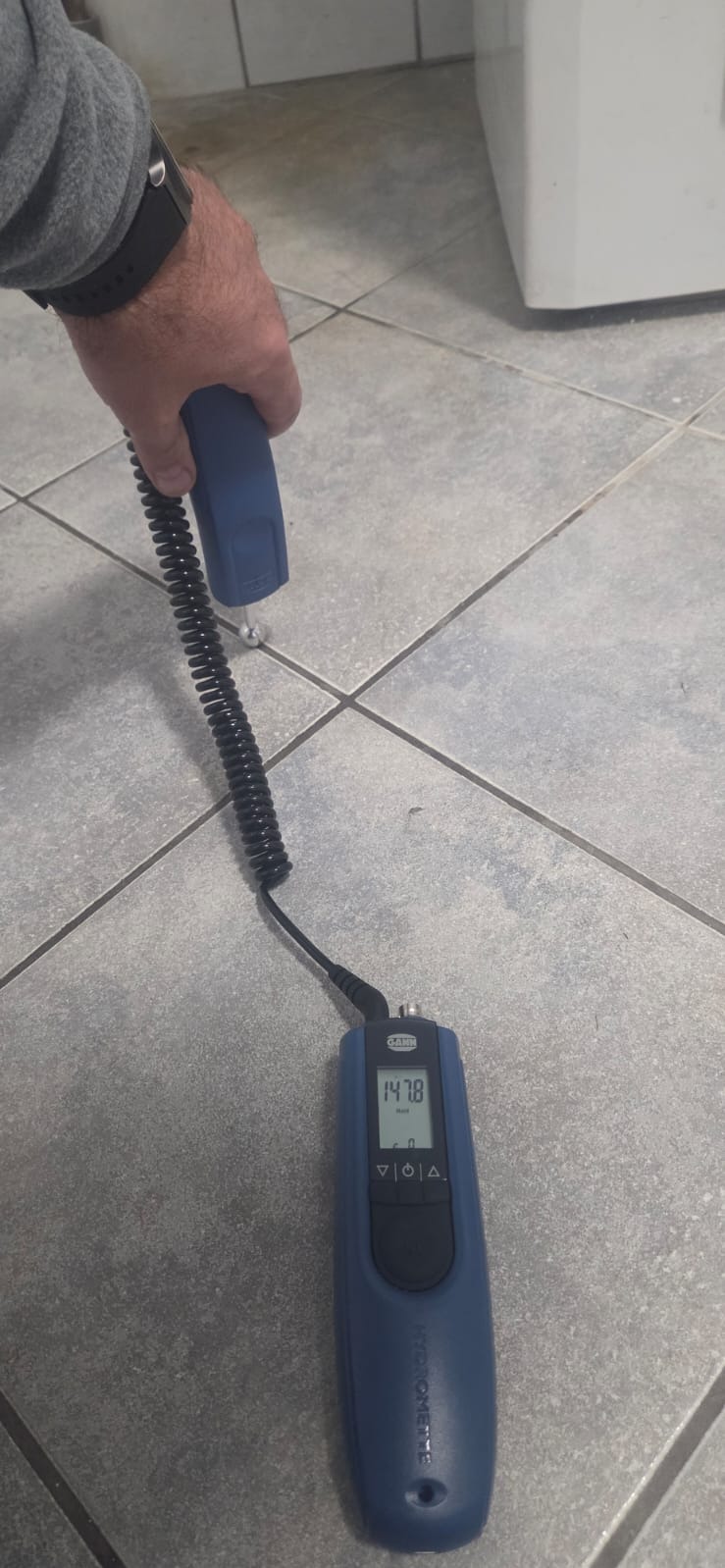 Pomiar wilgotności posadzki za pomocą wilgotnościomierza Gann, na szarych płytkach podłogowych. Widoczny fragment dłoni i urządzenia.