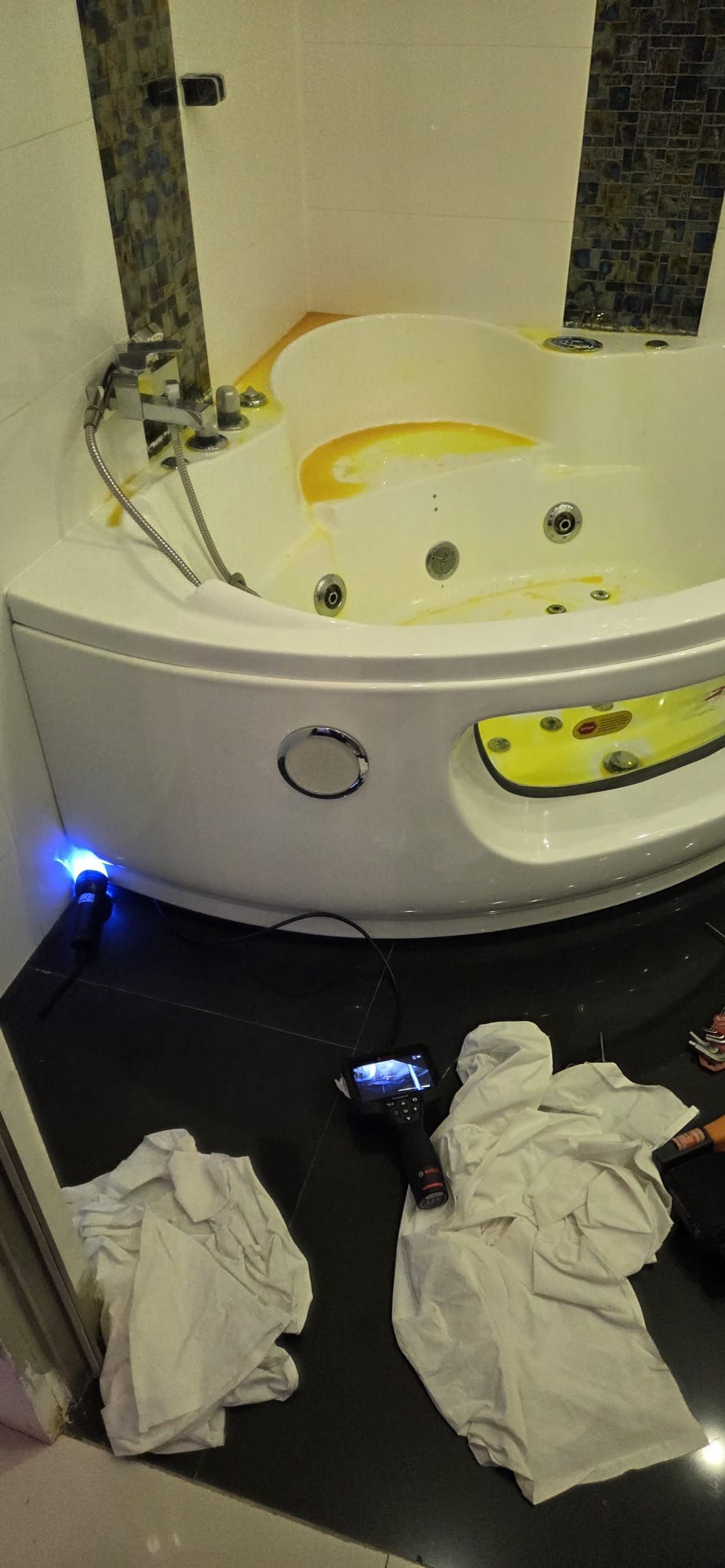 Biała wanna z hydromasażem wypełniona żółtą cieczą, inspekcja endoskopem z niebieskim światłem i ekranem. Widoczne narzędzia i szmatki na czarnej podłodze.