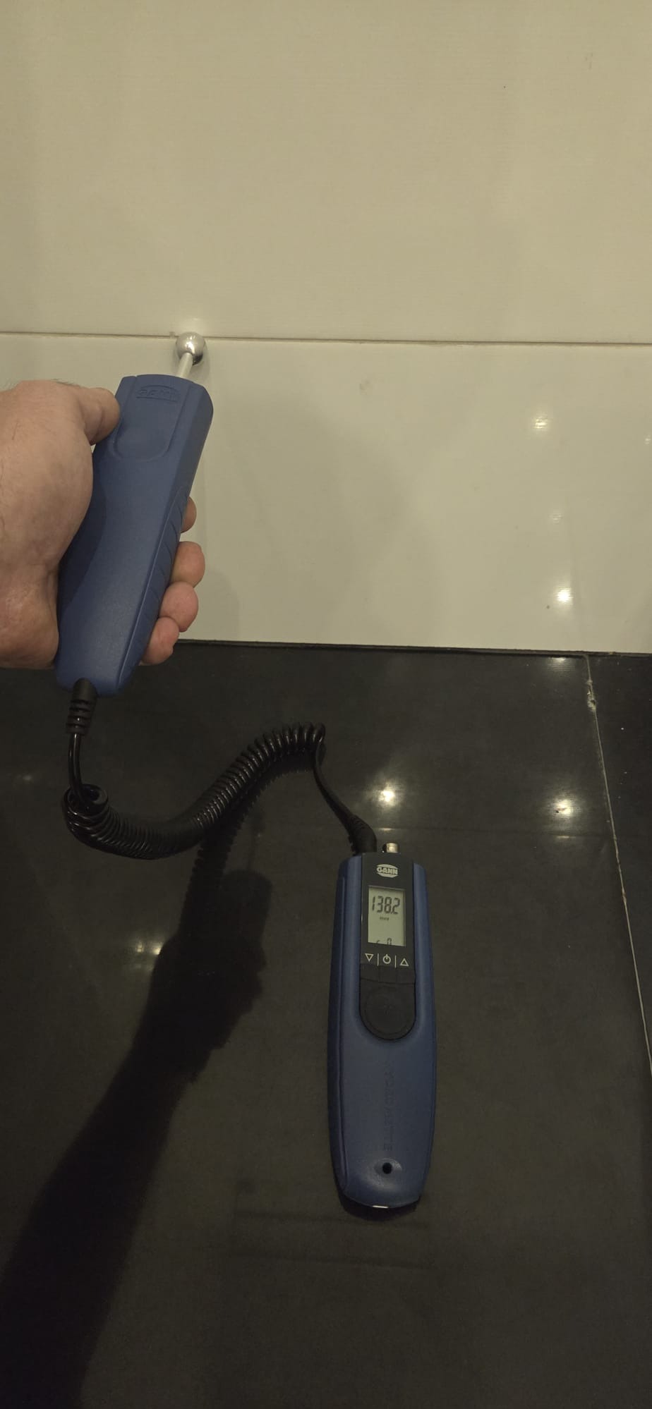 Pomiar wilgotności ściany za pomocą elektronicznego miernika wilgoci. Widoczny fragment ściany z połyskliwych płytek i czarna podłoga. Miernik w dłoni.