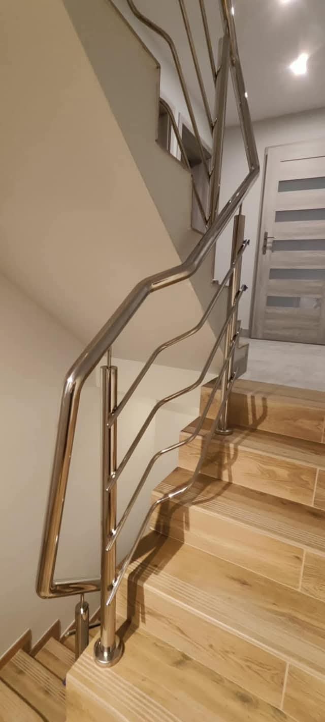 Nowoczesna, błyszcząca balustrada ze stali nierdzewnej o falistym wzorze, zamontowana przy schodach wyłożonych płytkami imitującymi drewno. Widok z dołu.