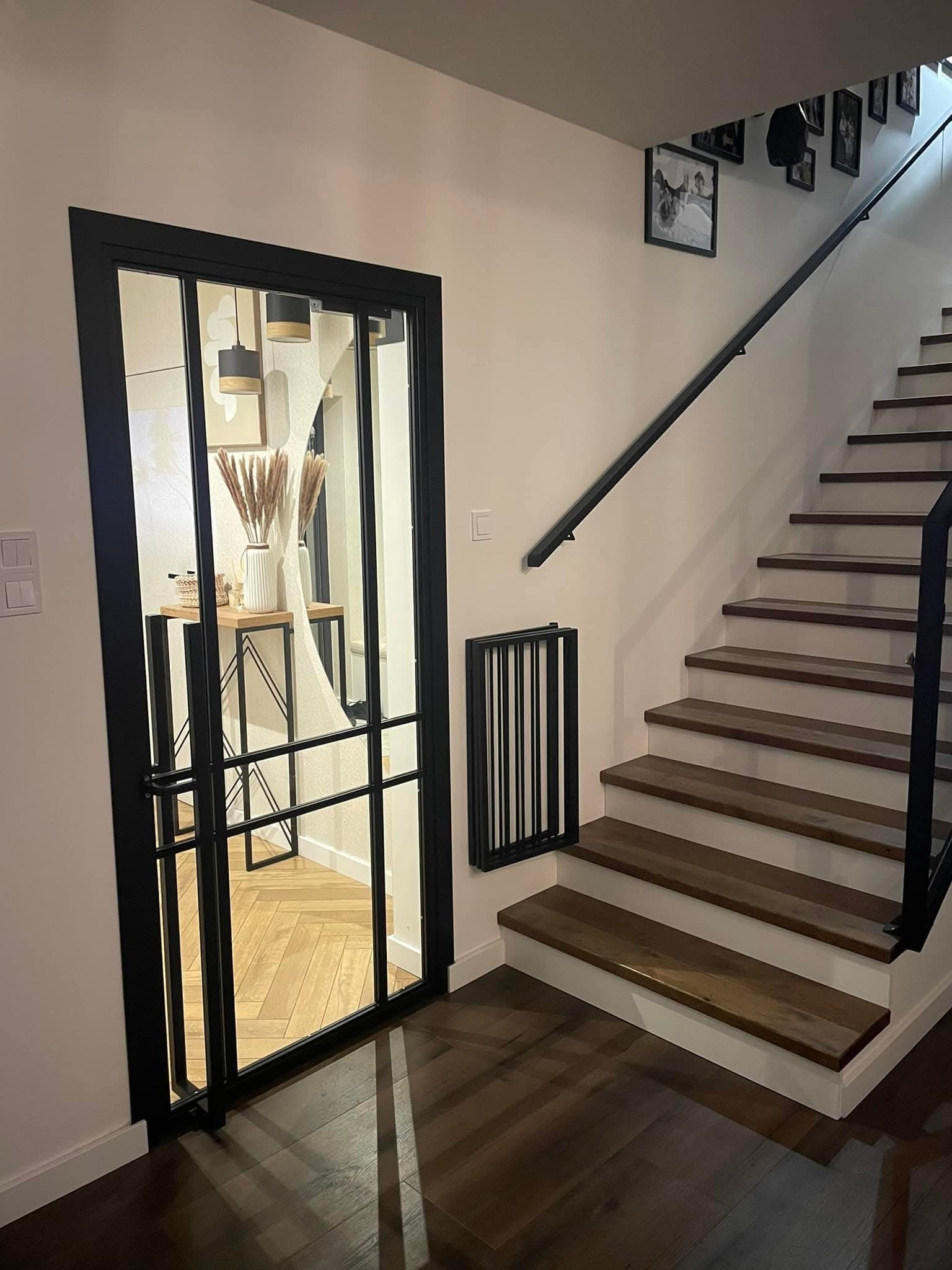 Nowoczesna, czarna, metalowa konstrukcja drzwi z przeszkleniami, oddzielająca przestrzeń. Widoczne schody z drewnianymi stopniami i czarną balustradą.