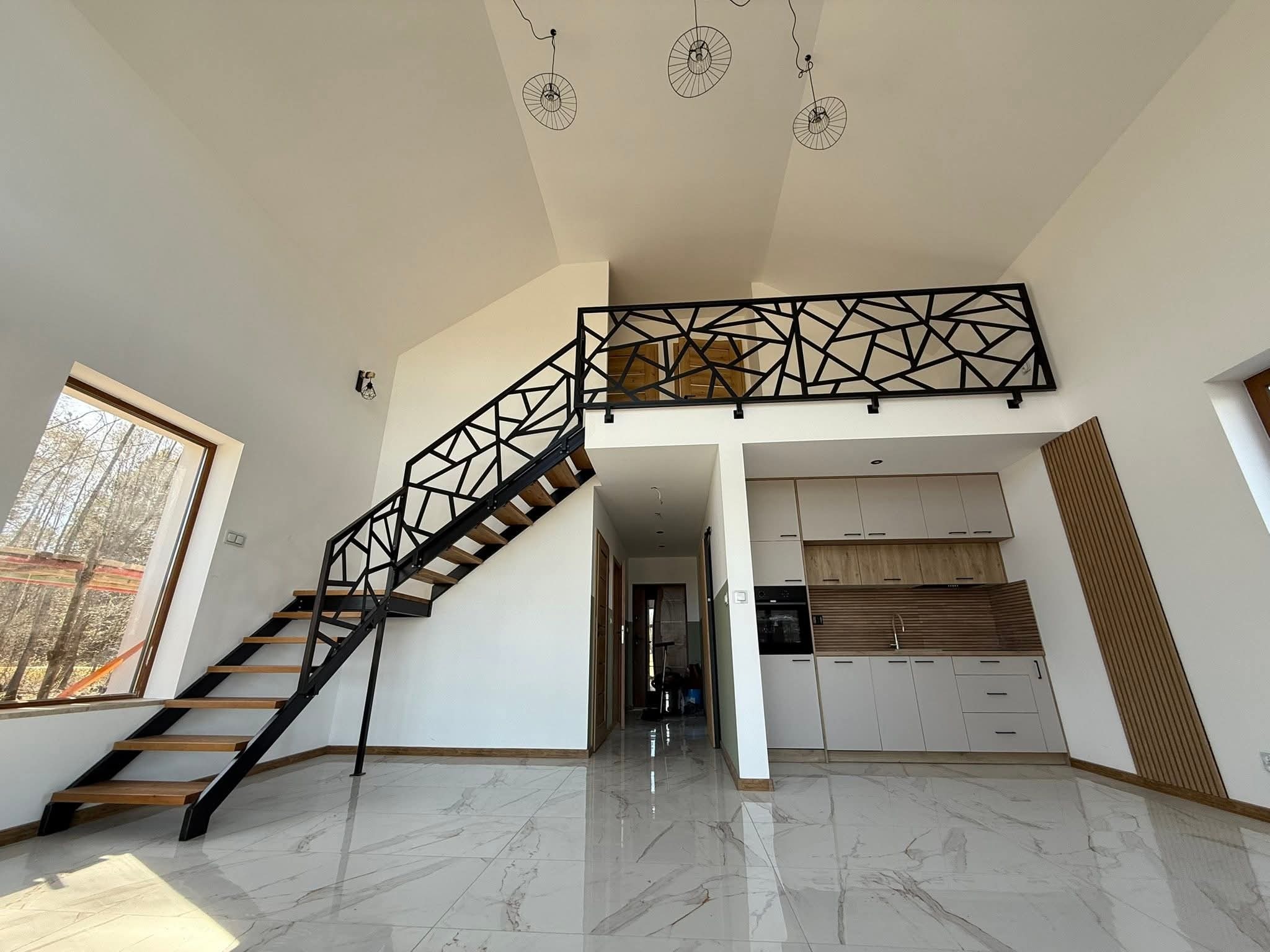 Nowoczesne schody z drewnianymi stopniami i czarną, geometryczną konstrukcją. Balustrada na antresoli z motywem abstrakcyjnym. Jasne wnętrze z marmurową podłogą.