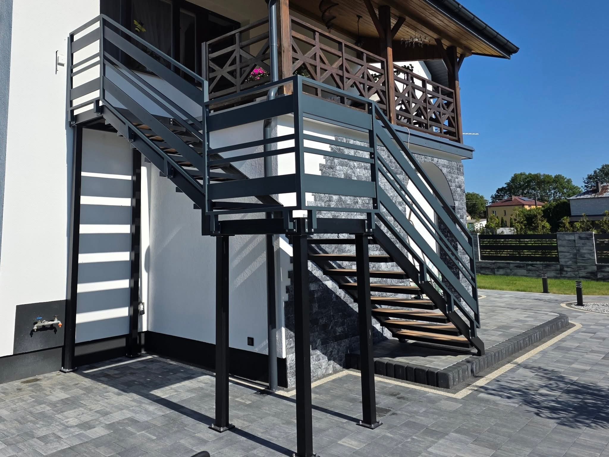 Zewnętrzne schody metalowe z szarą balustradą prowadzące na balkon z drewnianą konstrukcją. Nowoczesny design, kostka brukowa na podłożu, jasne tło budynku.