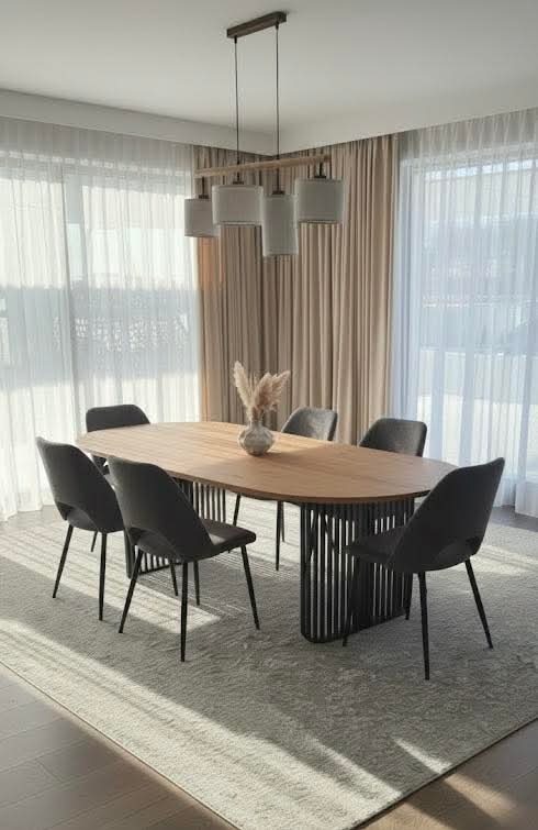 Nowoczesna jadalnia z owalnym stołem z drewnianym blatem i czarną, ażurową podstawą. Nad stołem wisi lampa z kilkoma abażurami. Całość w jasnej tonacji.