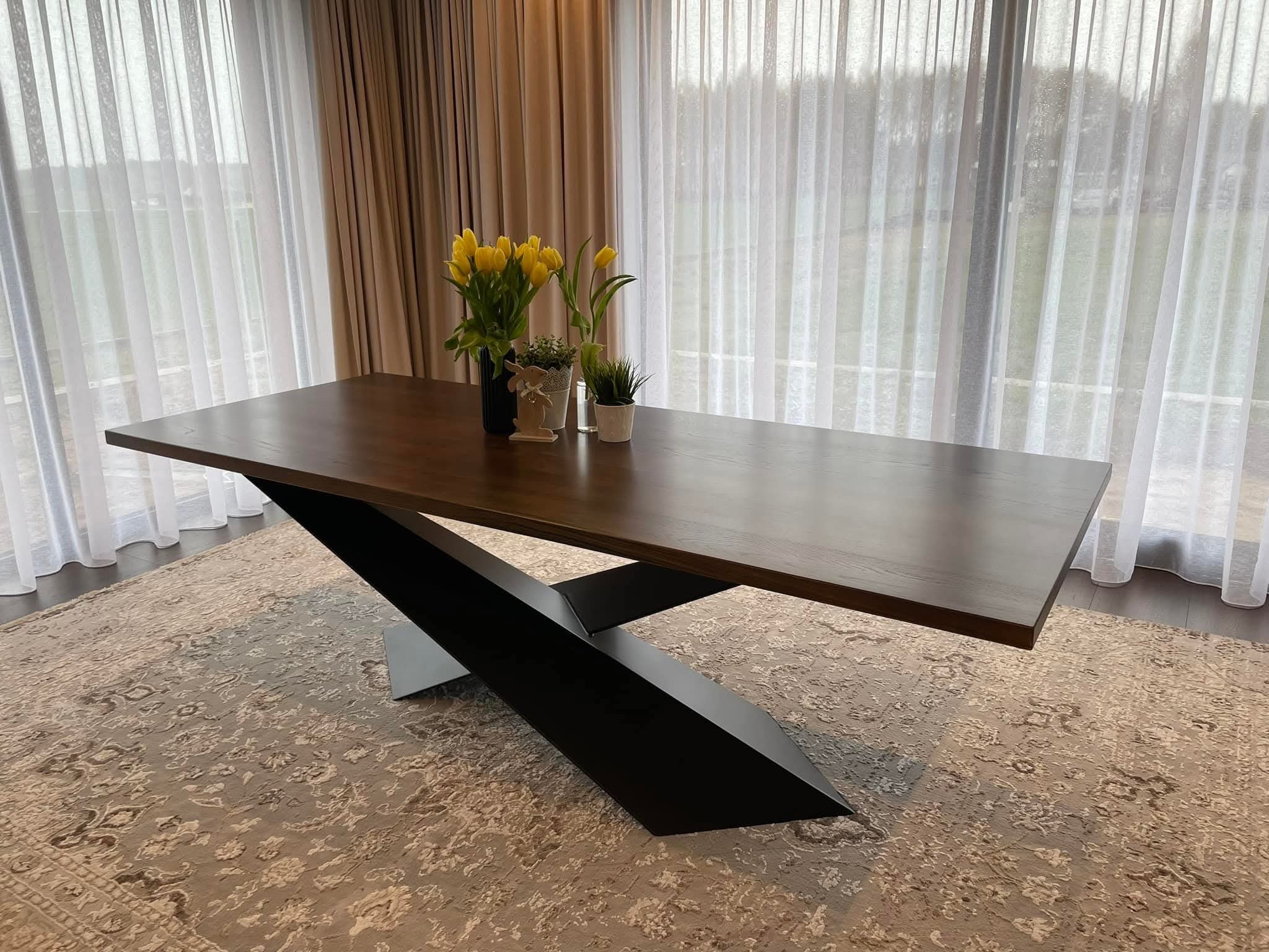 Stół z ciemnego drewna na metalowej, geometrycznej podstawie w nowoczesnym wnętrzu z zasłonami i dywanem. Na blacie dekoracje: kwiaty i figurka.