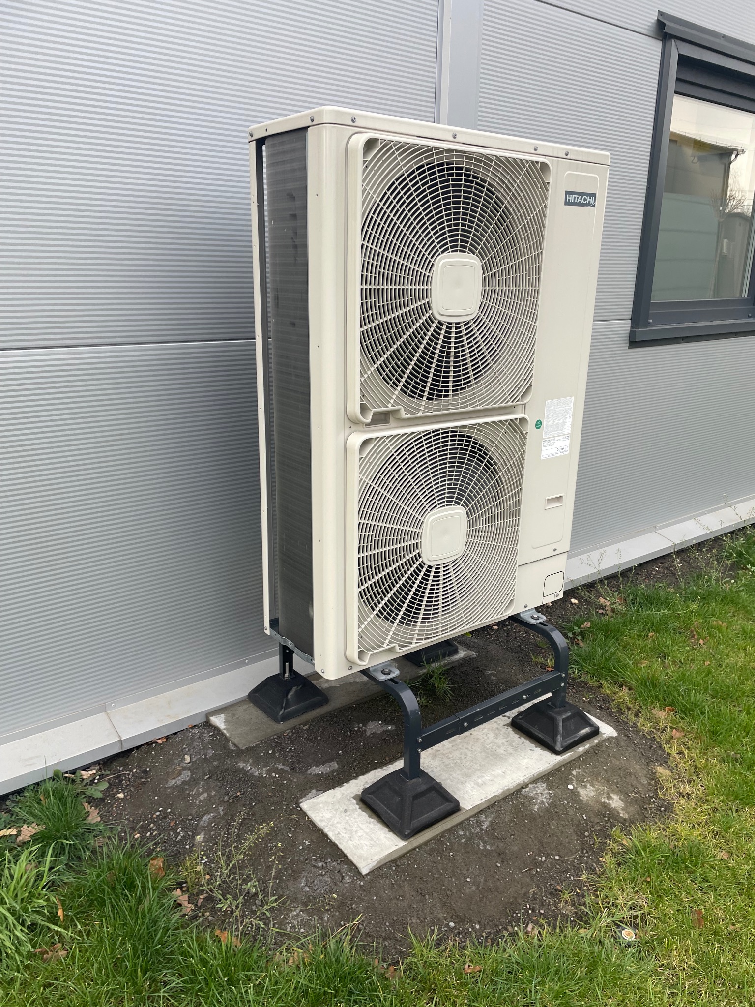 Zewnętrzna jednostka klimatyzacji Hitachi, zamontowana na metalowej konstrukcji przy budynku w Częstochowie. Widoczne dwa wentylatory i szara elewacja budynku.