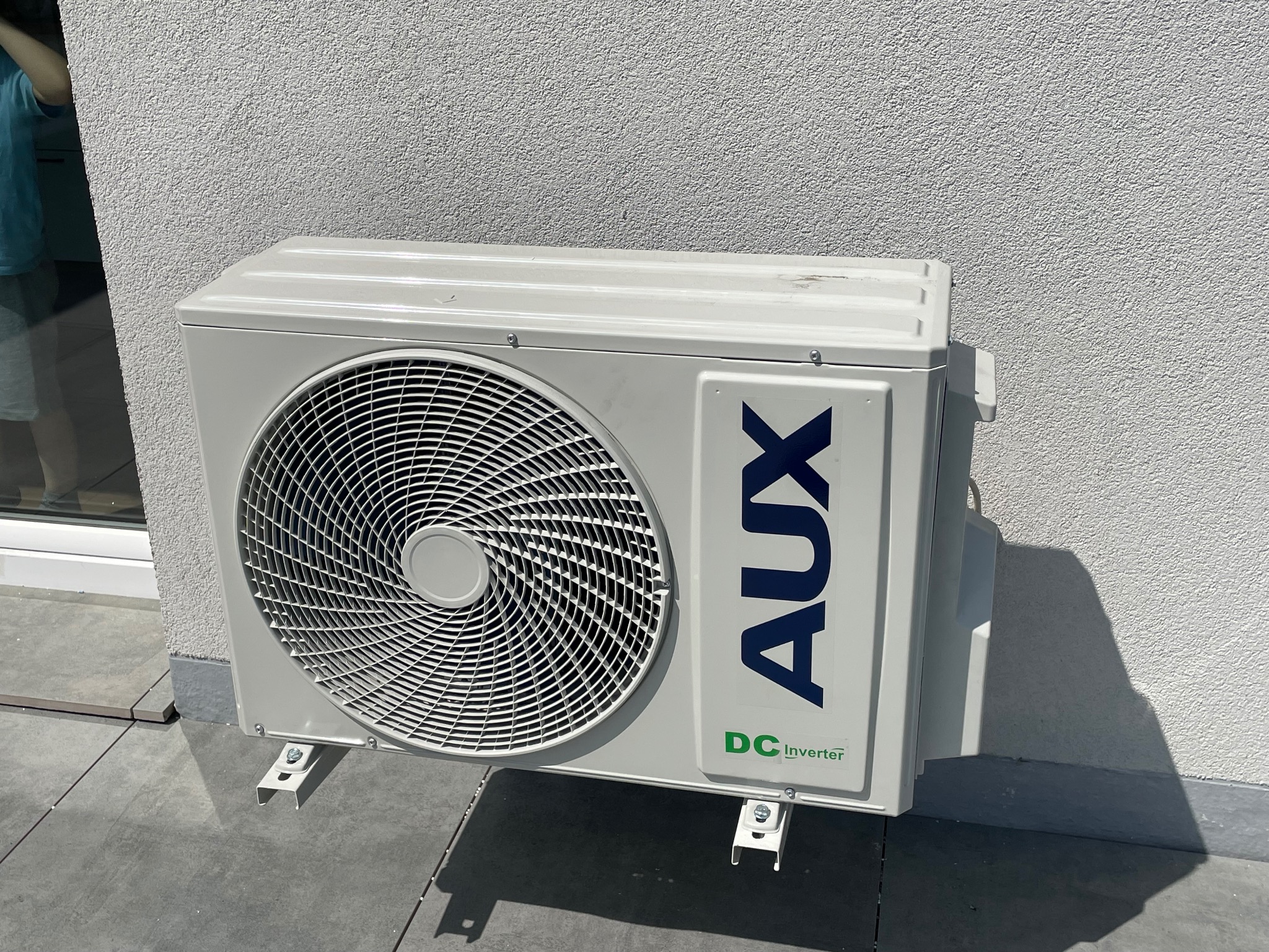 Biała jednostka zewnętrzna klimatyzacji AUX DC Inverter zamontowana na szarym podłożu przy teksturowanej, jasnej ścianie budynku. Widoczne śruby mocujące.