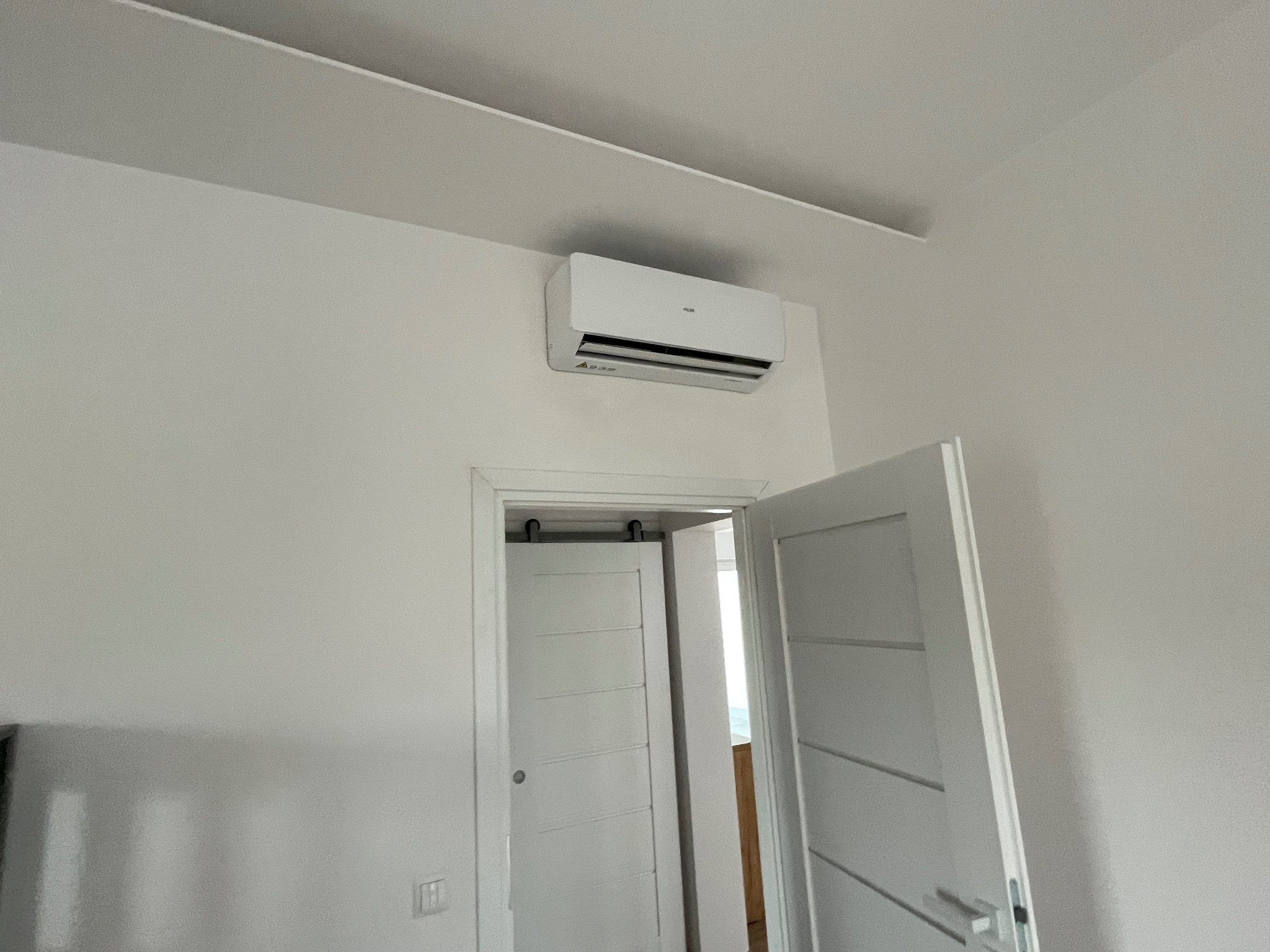 Biały klimatyzator AUX zamontowany na ścianie nad otwartymi, przesuwnymi drzwiami w jasnym pomieszczeniu. Minimalistyczny design, neutralne tło.
