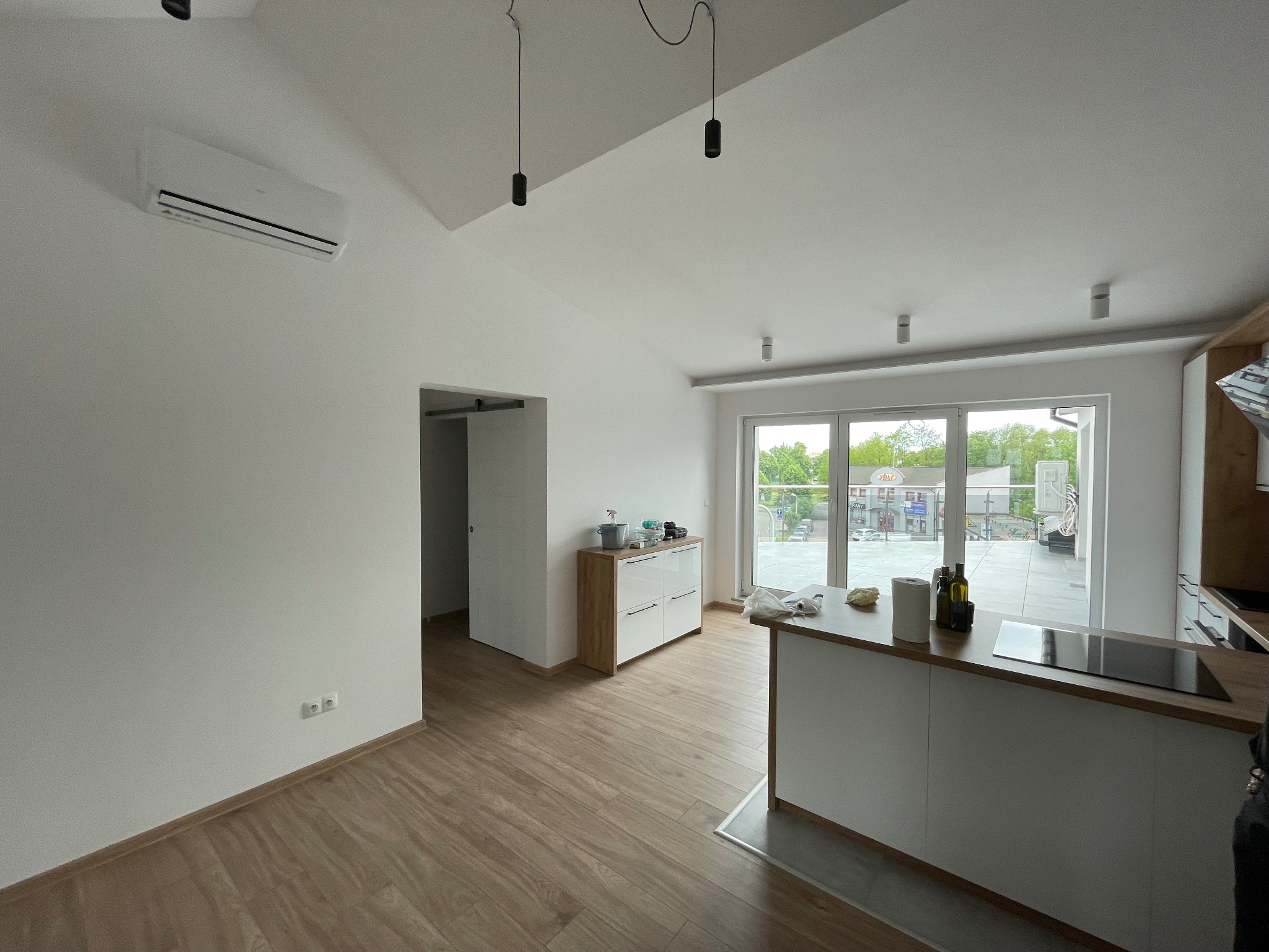 Nowoczesne wnętrze z zamontowaną klimatyzacją na ścianie. Widoczne minimalistyczne meble, drewniana podłoga i duże okna z widokiem na zewnątrz. Białe ściany i sufit.