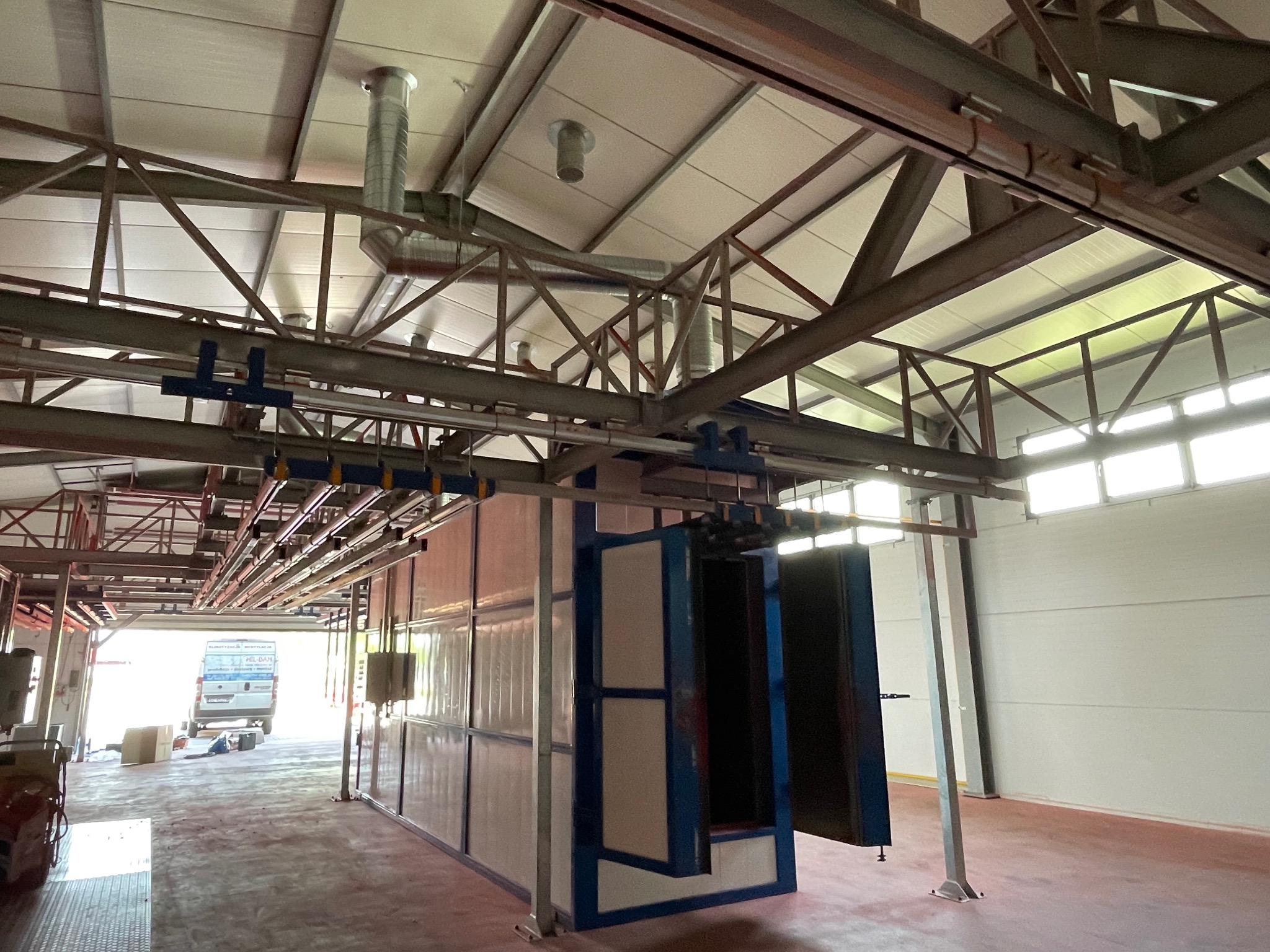 Wnętrze hali z systemem wentylacji pod stropem i kabiną lakierniczą z otwartymi drzwiami. Widoczny wjazd do hali i zaparkowany samochód dostawczy.