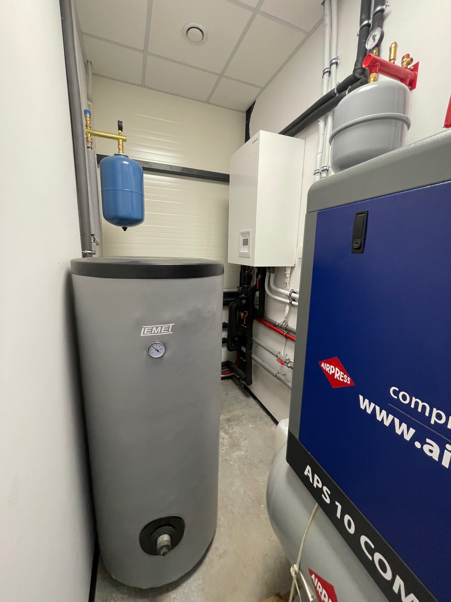 Zestaw urządzeń w kotłowni: szary zbiornik EMEI z manometrem, biały piec, niebieski zbiornik wyrównawczy i kompresor Airpress APS 10 COM z widocznym adresem strony.