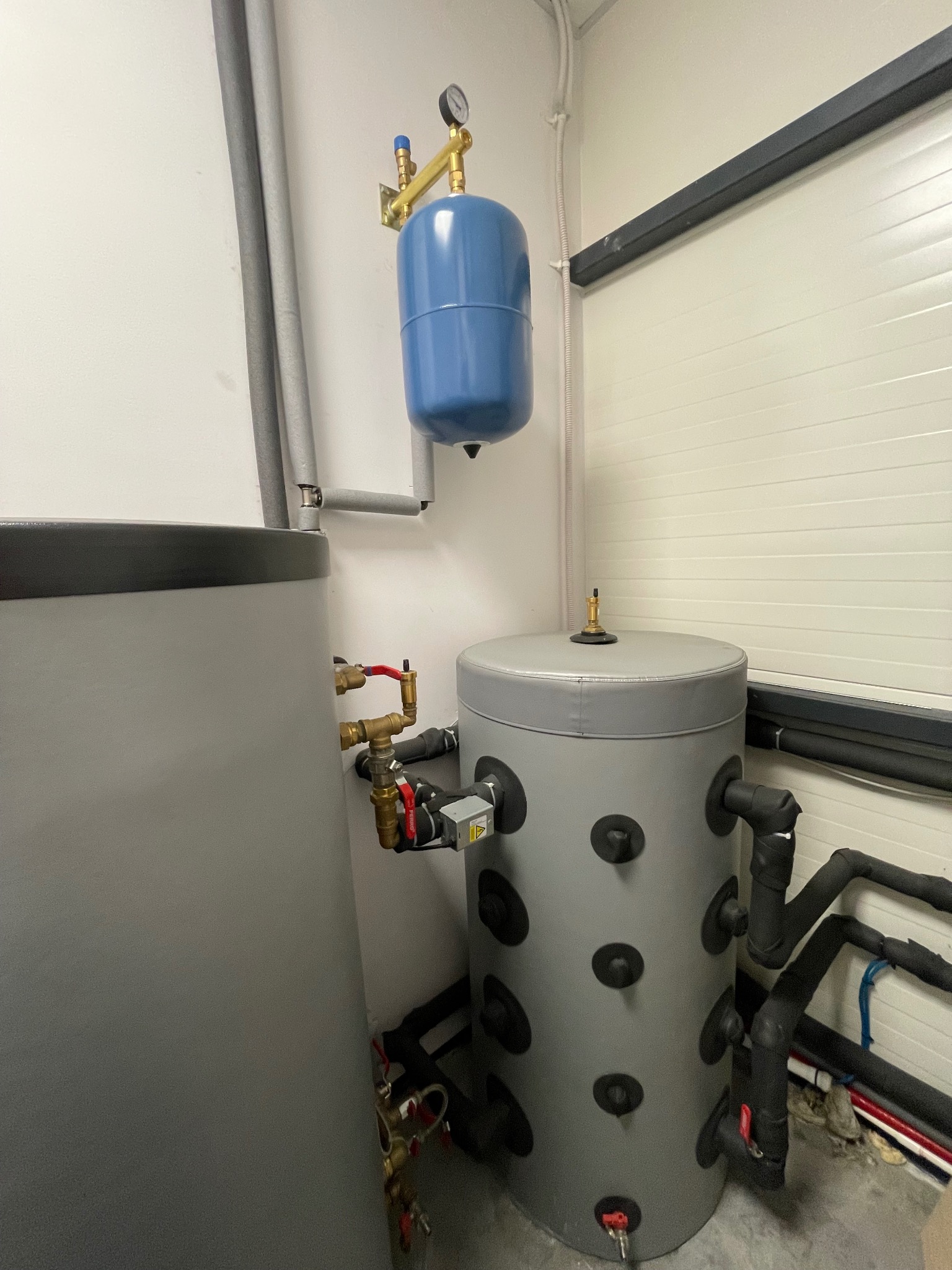 Szary zbiornik buforowy z izolacją i niebieski zbiornik wyrównawczy z manometrem, połączone rurami w kotłowni. Widoczne zawory i izolacja termiczna rur.