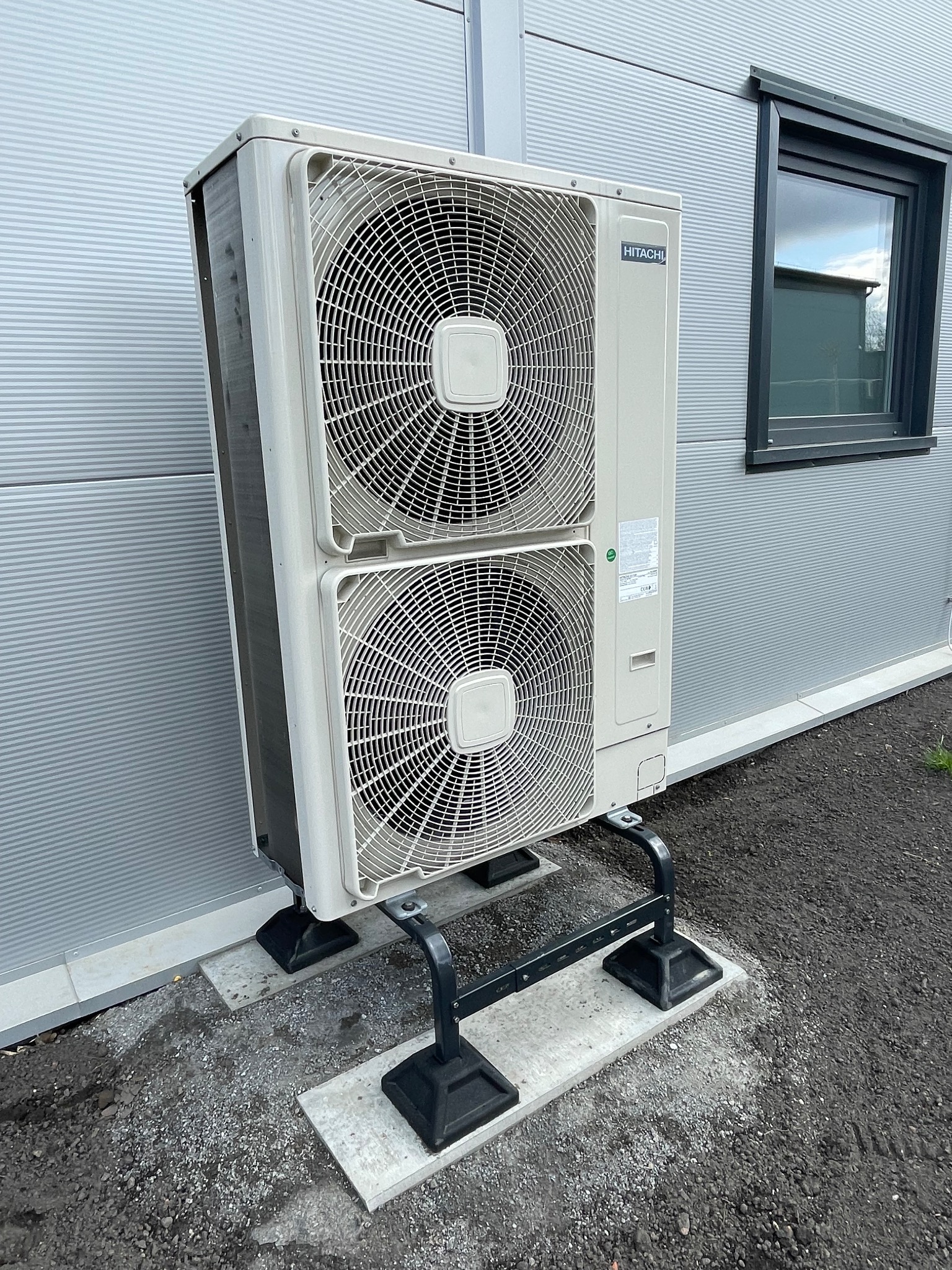 Zewnętrzna jednostka klimatyzacji Hitachi, zamontowana na metalowym stelażu na betonowych płytach, przy szarej ścianie budynku z oknem. Widok z dołu.