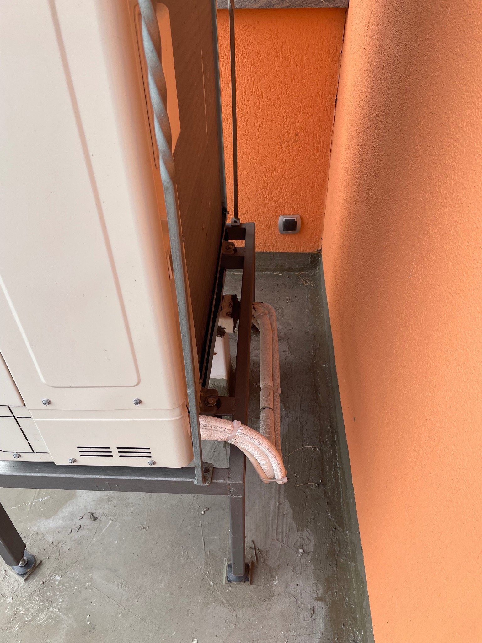 Zewnętrzna jednostka klimatyzacji na metalowej konstrukcji, z widocznymi rurami w izolacji, umieszczona przy pomarańczowej ścianie budynku. Widok z boku.