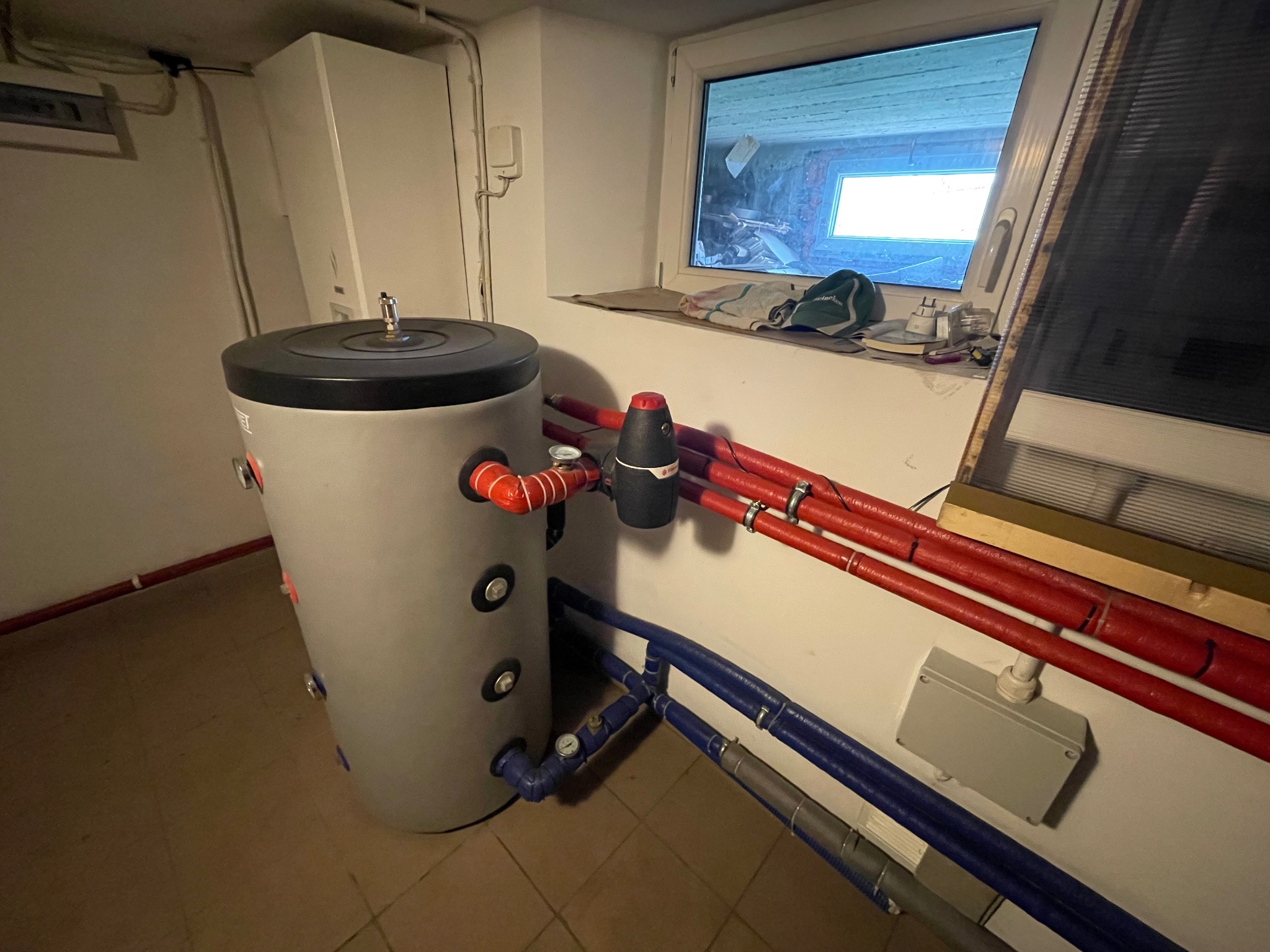 Szary zbiornik buforowy z czerwonymi i niebieskimi rurami w piwnicy, widoczne okno z bałaganem na parapecie, instalacja grzewcza w pomieszczeniu technicznym.