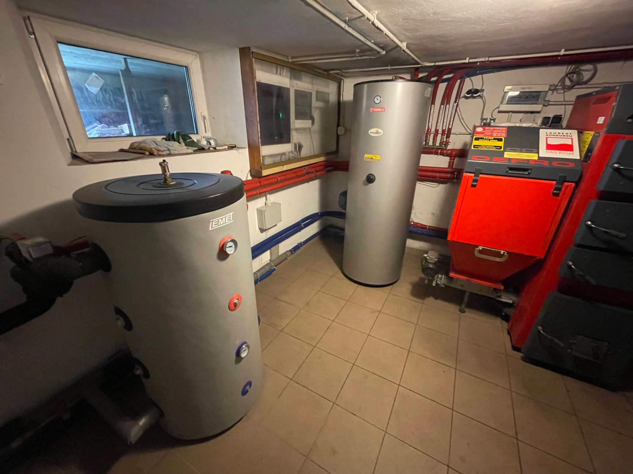 Kotłownia z dwoma szarymi zbiornikami EME i Termica, czerwonym kotłem Defro, rurami i oknem. Widok na instalację w piwnicy z terakotą.