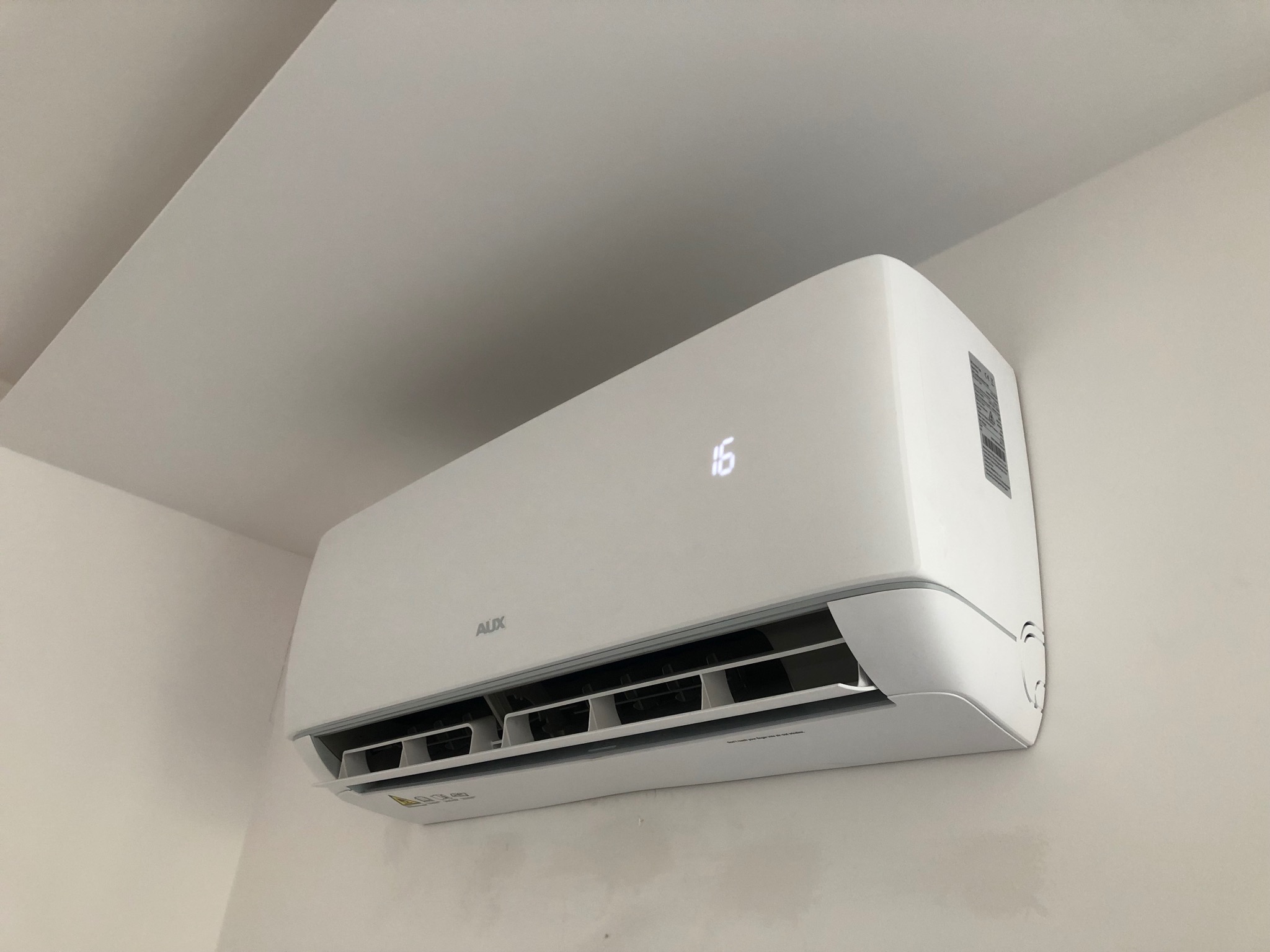 Biała klimatyzacja AUX zamontowana na ścianie, widoczny wyświetlacz z temperaturą 16 stopni, minimalistyczny design, jasne wnętrze.