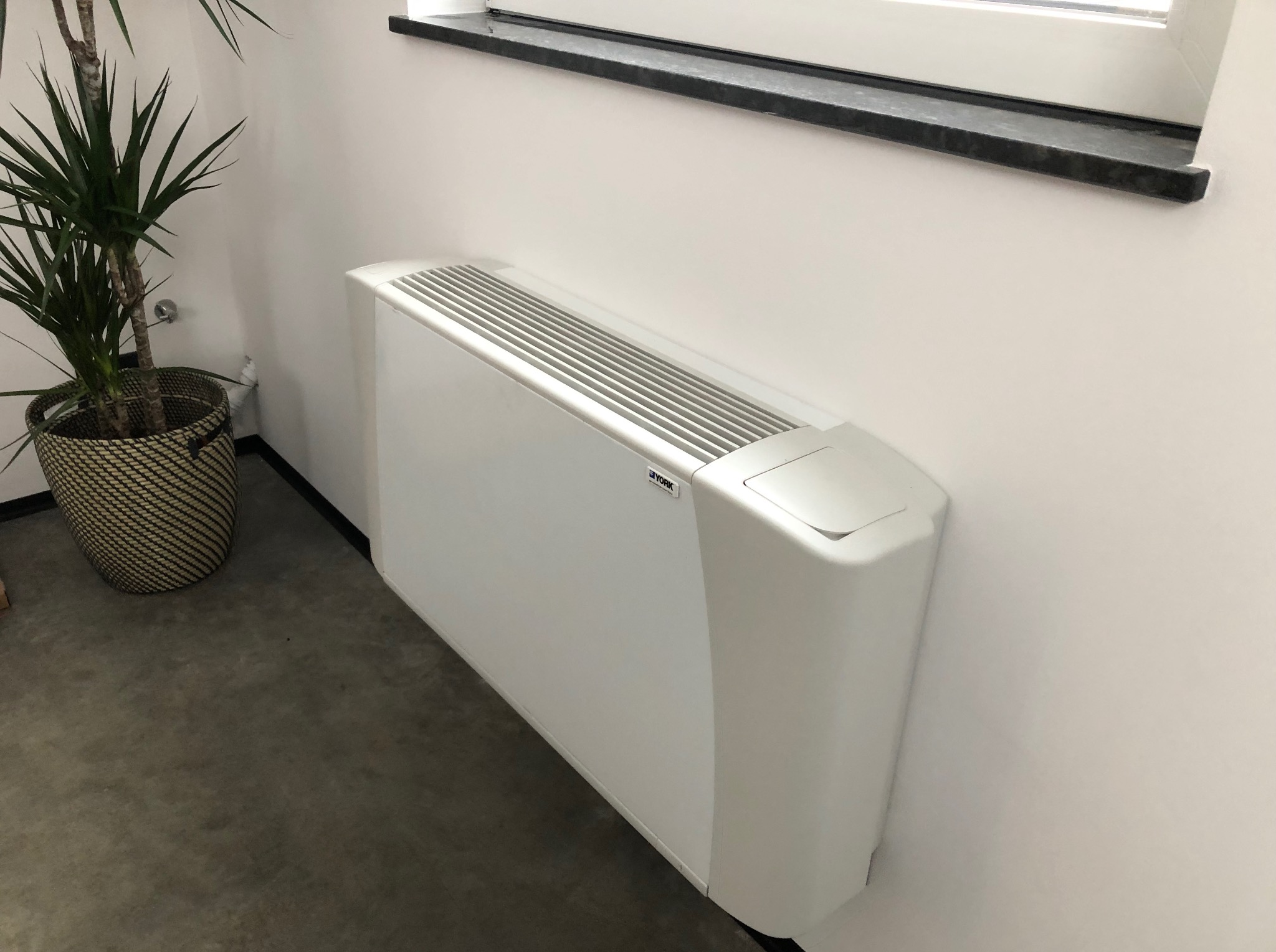 Biały klimakonwektor pod oknem, w rogu pomieszczenia, obok doniczka z zieloną rośliną. Minimalistyczny design, szara podłoga, czarny parapet.