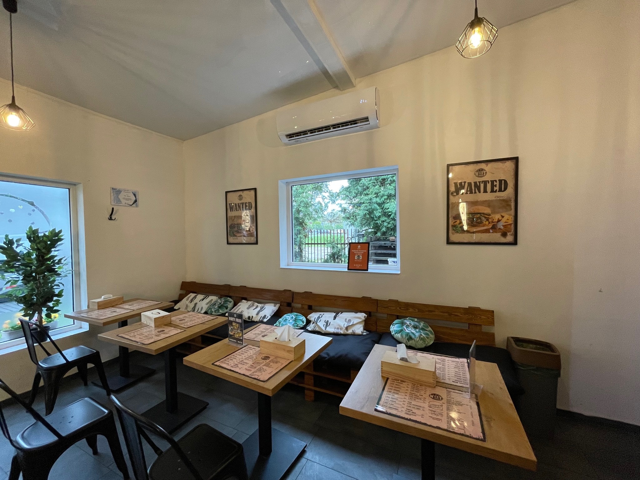 Klimatyzator ścienny w jasnym wnętrzu restauracji. Widoczne stoliki, krzesła, okno z zielenią i plakaty na ścianach. Oświetlenie punktowe.