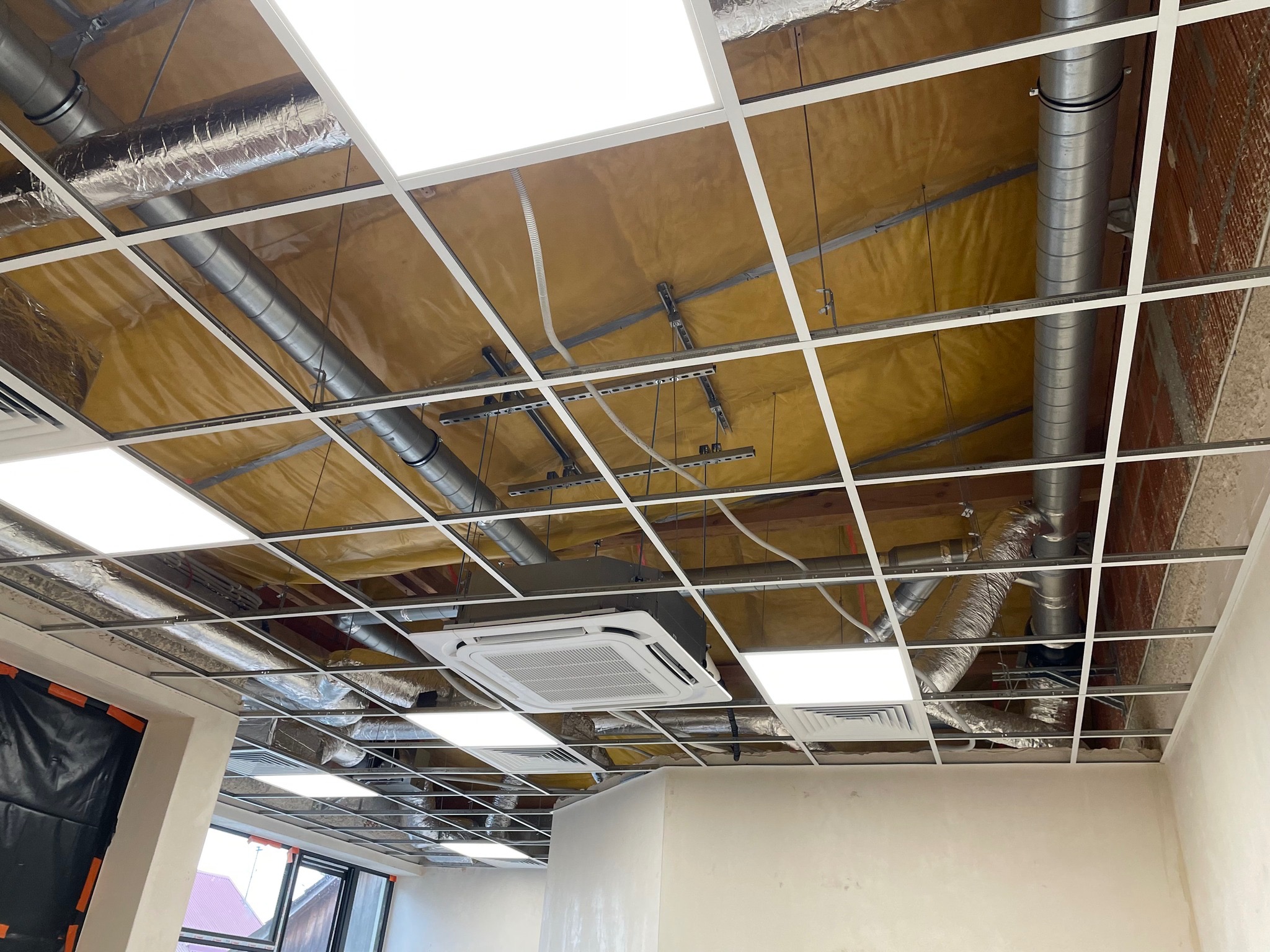 Sufit podwieszany z zamontowaną kasetą klimatyzacyjną, widoczne rury wentylacyjne i instalacja elektryczna. Żółta izolacja termiczna w tle.