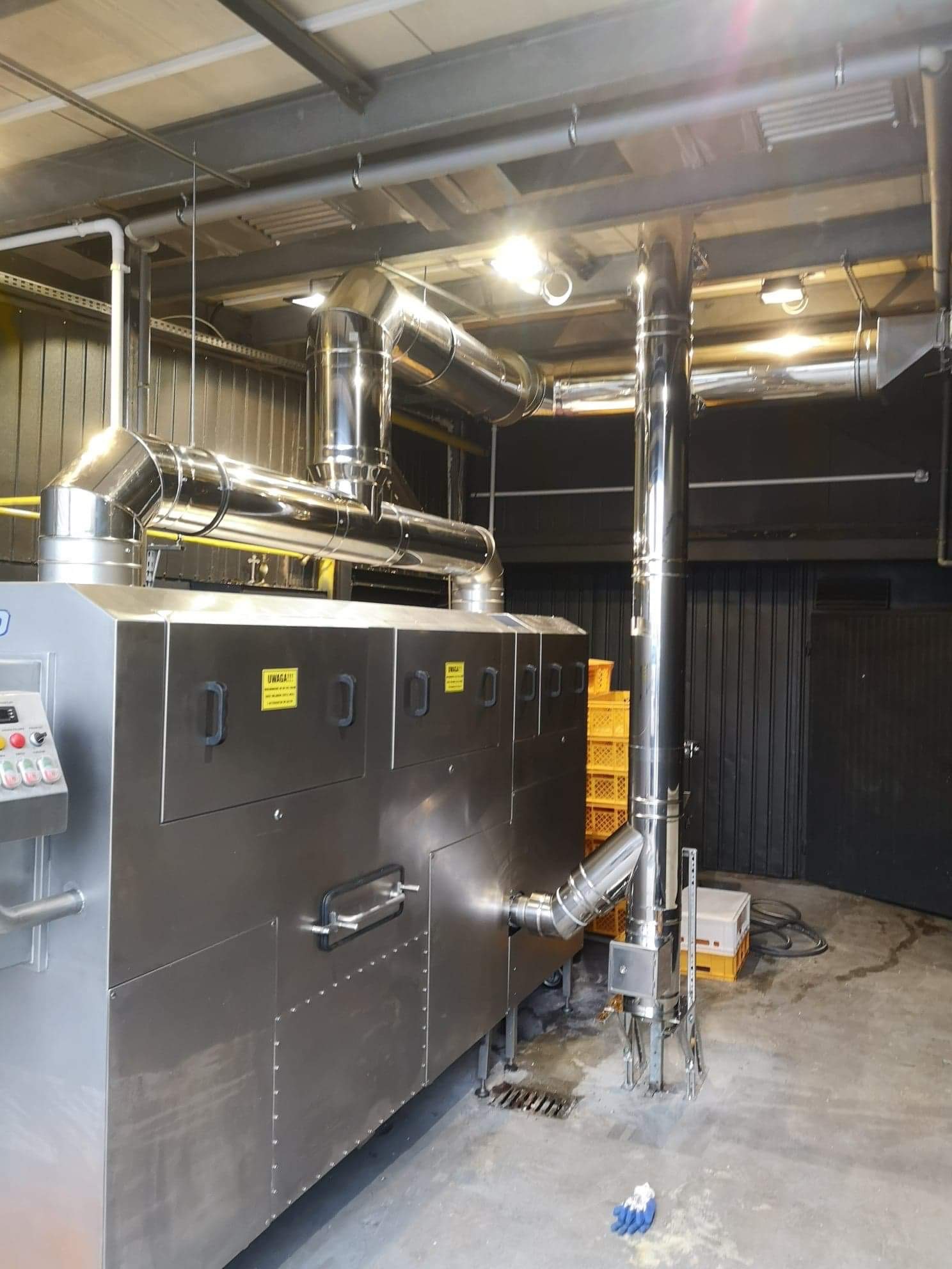 Kompleksowa instalacja wentylacyjna z rurami ze stali nierdzewnej w hali przemysłowej. Widoczne elementy sterujące i żółte skrzynki w tle.