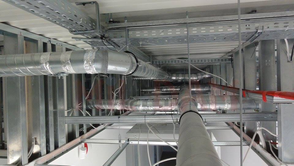 Srebrne rury wentylacyjne pod sufitem, widok z dołu. Metalowe konstrukcje wsporcze i kable. Instalacja w trakcie montażu, widoczne łączenia rur.