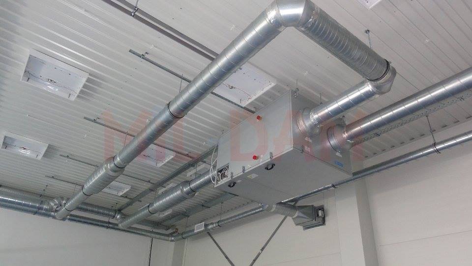 Srebrne rury wentylacyjne i centrala rekuperacyjna zamontowane pod sufitem w hali. Widoczne elementy mocujące i oświetlenie sufitowe.