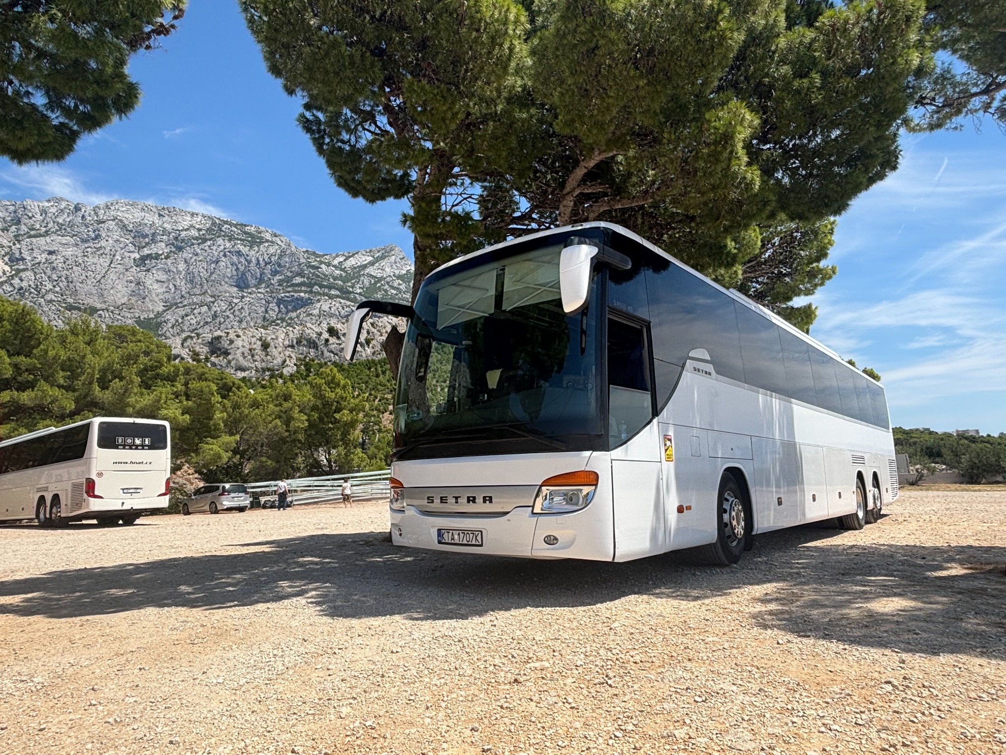 Biały autokar Setra na żwirowym parkingu, z otwartymi drzwiami, w tle góry i drzewa. Drugi autokar widoczny w oddali. Słoneczny dzień.