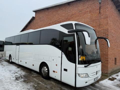 Biały autokar Mercedes-Benz na tle ceglanego budynku. Widoczny znak ostrzegawczy z symbolem dzieci. Zimowa sceneria, lekko ośnieżone podłoże. Profesjonalny transport.