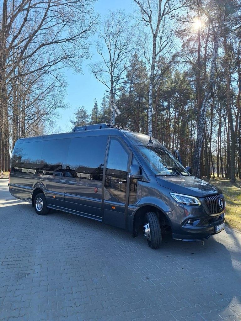 Elegancki, grafitowy bus Mercedes-Benz na tle drzew i kostki brukowej. Widoczny z boku, z detalami jak relingi dachowe i chromowane elementy. Słoneczny dzień.