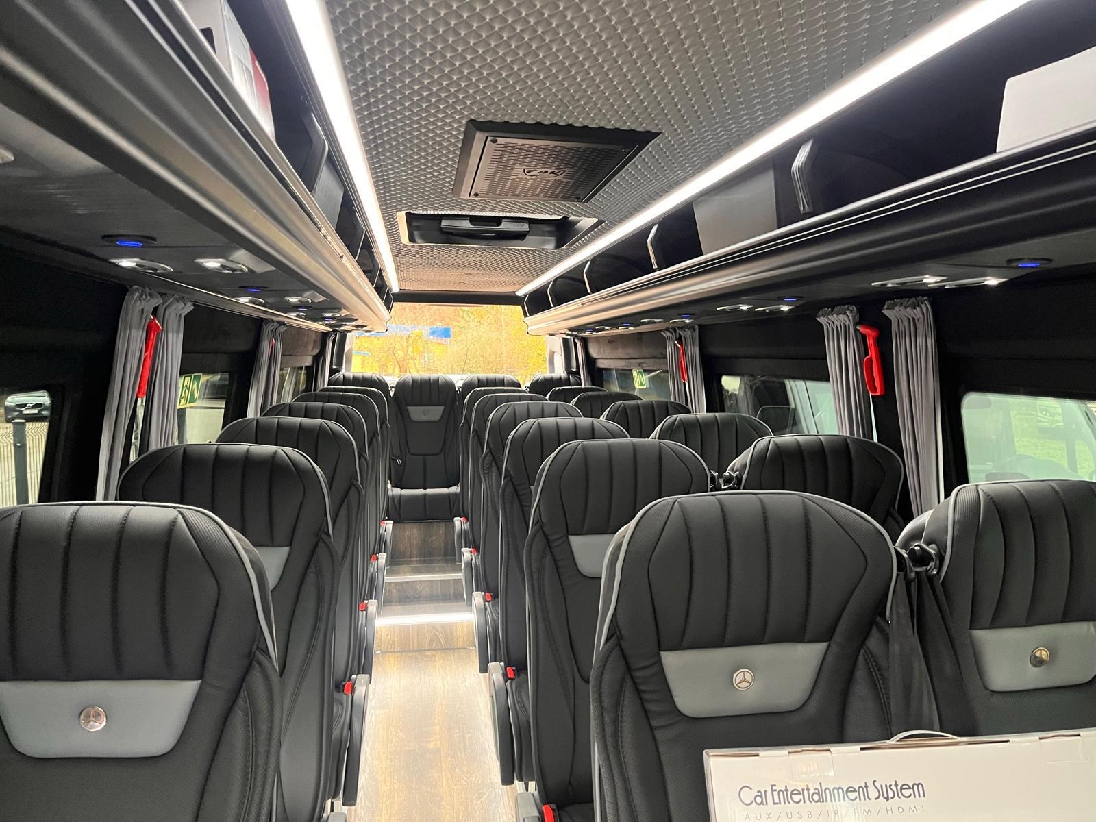 Wnętrze luksusowego busa z czarnymi skórzanymi fotelami z szarymi wstawkami, oświetleniem LED i zasłonami w oknach. Widok na schodki i tylne siedzenia.