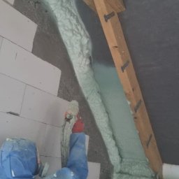 Pur-Up Docieplenia pianą PUR Pomorskie - Ocieplanie poddasza pianką PUR: pracownik w kombinezonie aplikuje pianę między krokwiami i ścianą z bloczków. Widoczna struktura piany i drewniane elementy konstrukcji dachu.
