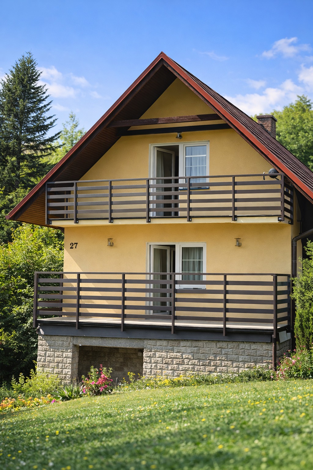 Dwukondygnacyjny dom z żółtą elewacją i balkonami z poziomymi, brązowymi barierkami. Widok na zadbany ogród i błękitne niebo.