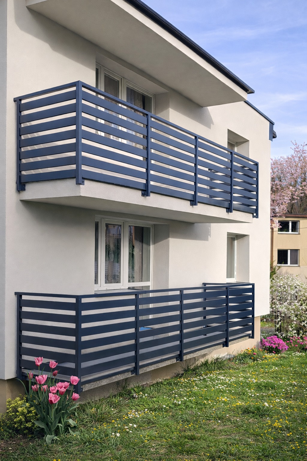 Dwukondygnacyjny budynek z balkonami zabezpieczonymi nowoczesnymi, poziomymi balustradami w kolorze grafitowym. W tle kwitnące drzewa i trawnik z tulipanami.