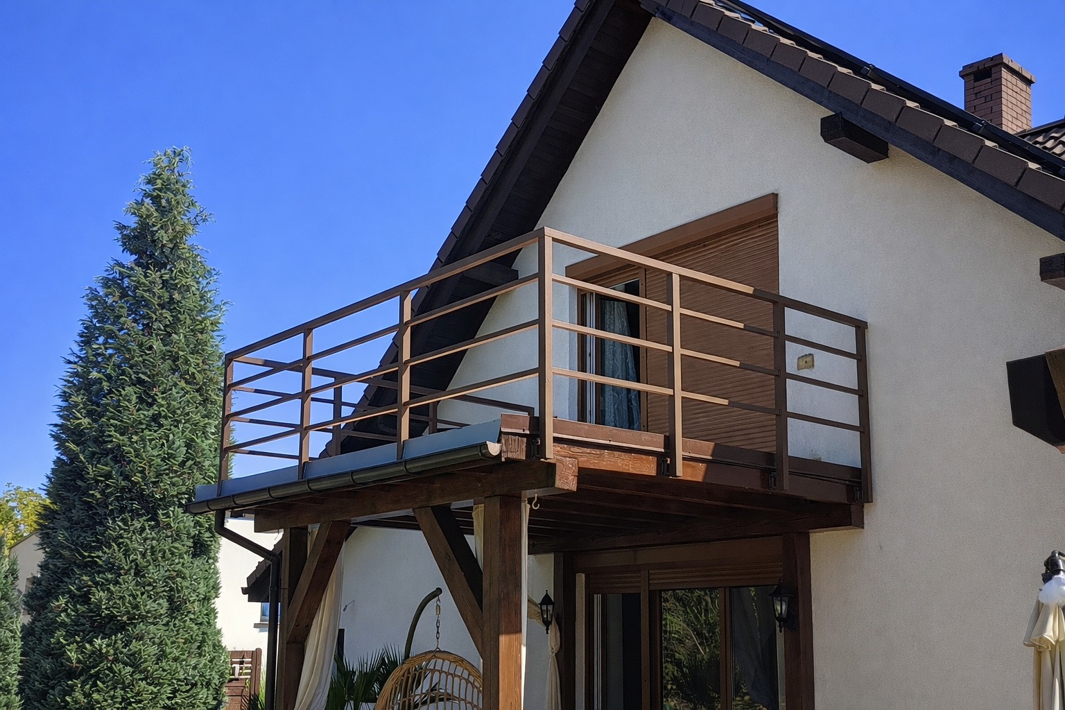 Balkon z brązową, metalową balustradą na tle jasnej elewacji domu i błękitnego nieba. Widoczny fragment ogrodu z wysokim drzewem iglastym.