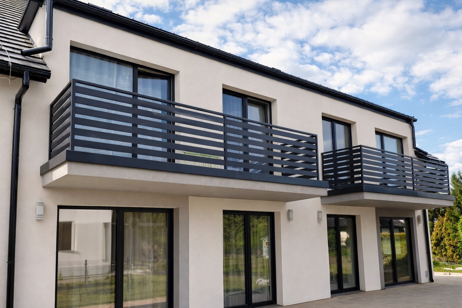 Nowoczesny budynek z balkonami zabezpieczonymi poziomymi, czarnymi balustradami. Jasna elewacja, ciemne ramy okien i drzwi. Błękitne niebo w tle.