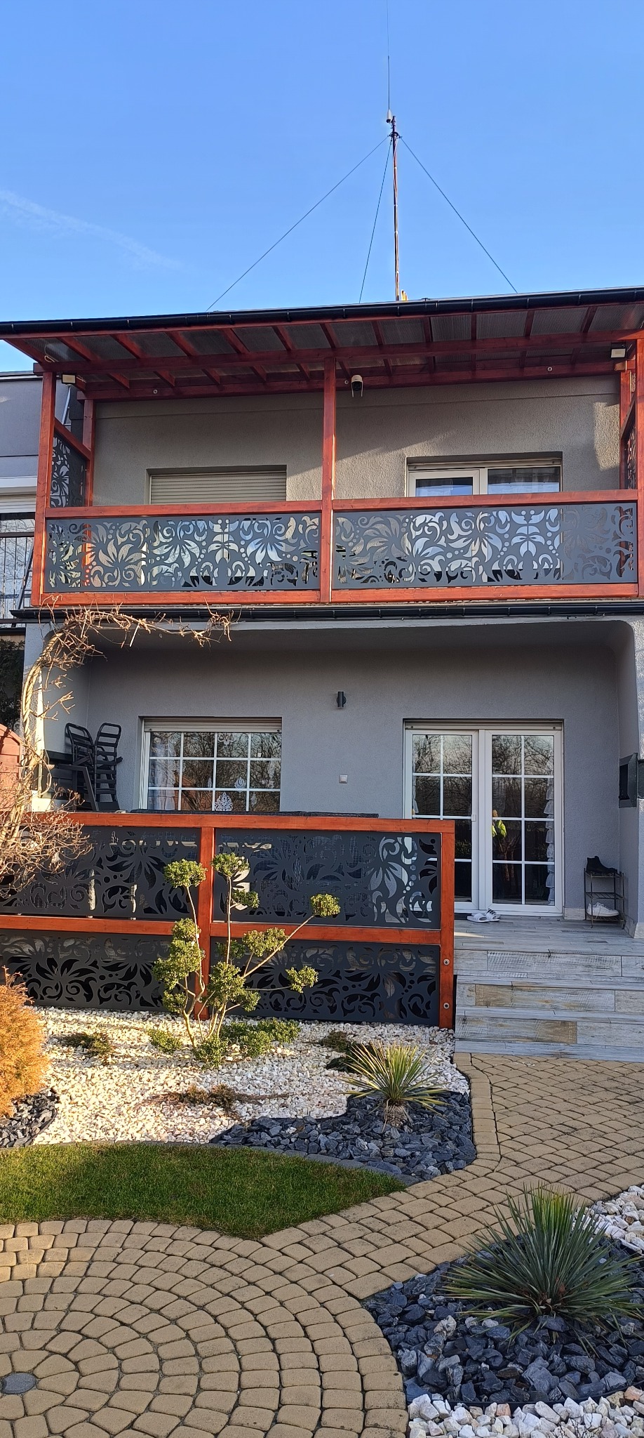 Dwukondygnacyjny dom z dekoracyjnymi, ażurowymi balustradami w kolorze grafitowym i drewnianą ramą. Balustrady na balkonie i przy tarasie.