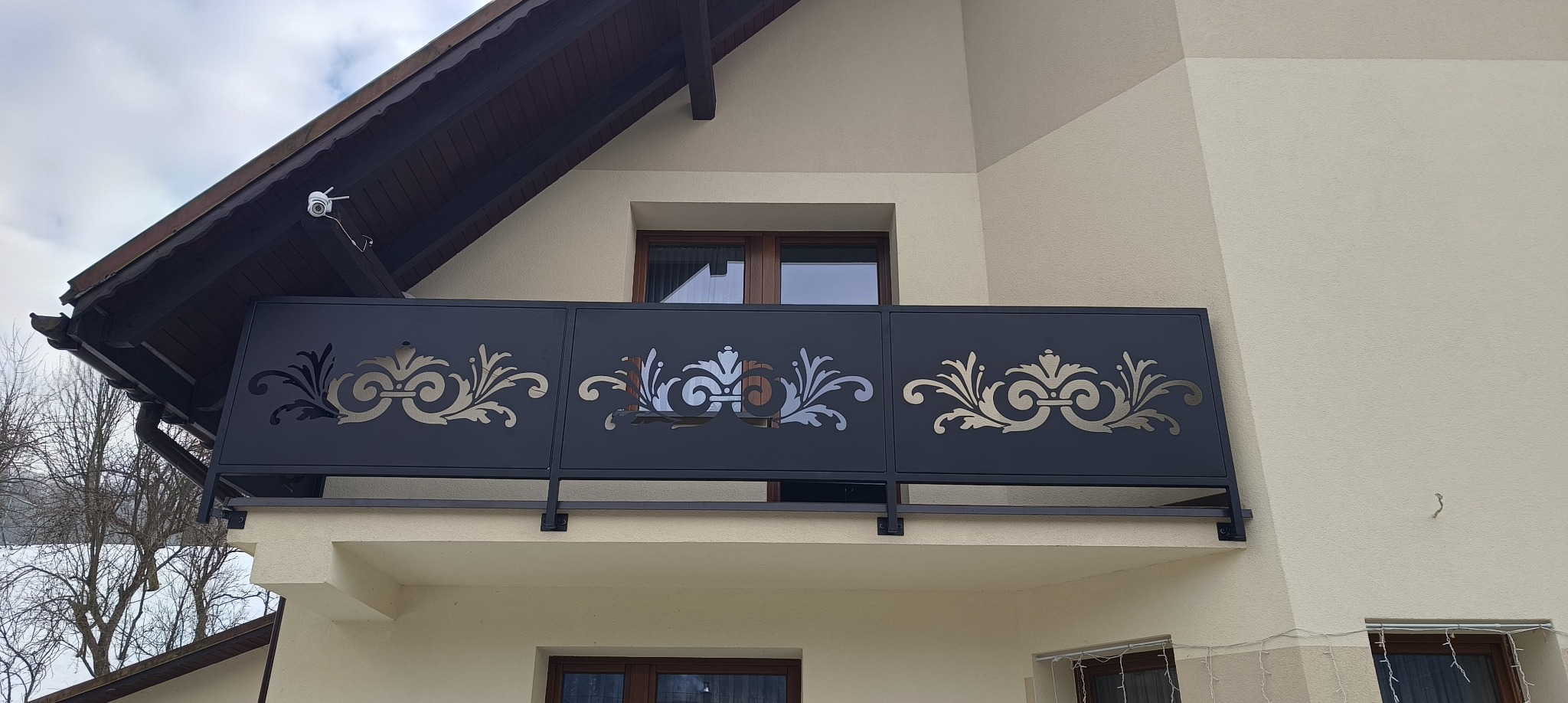 Czarna, metalowa balustrada balkonowa z ażurowym, roślinnym wzorem na tle elewacji domu i fragmentu dachu. Ujęcie z dołu.