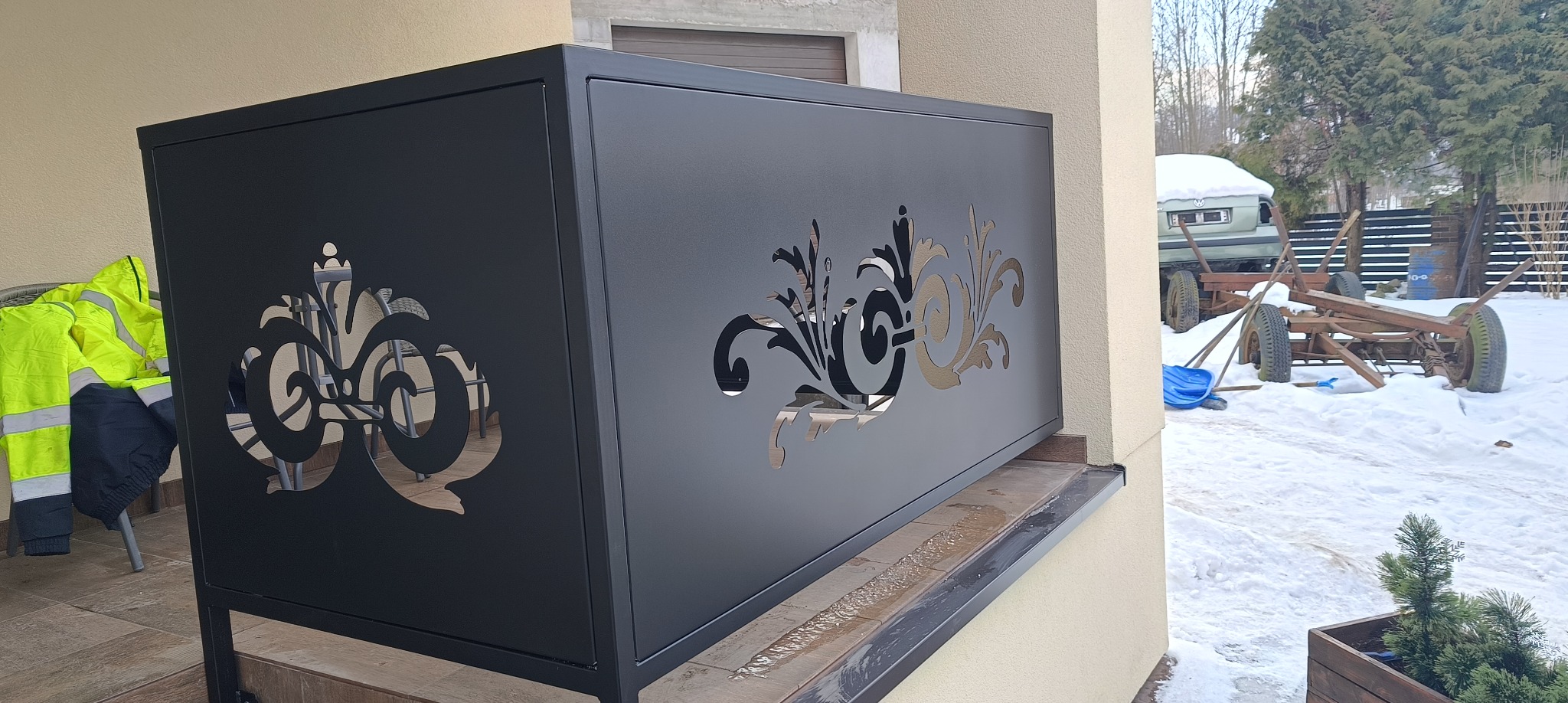 Czarna, metalowa balustrada balkonowa z wycinanymi, ozdobnymi wzorami, zamontowana na zewnątrz budynku. W tle widoczny ośnieżony krajobraz.