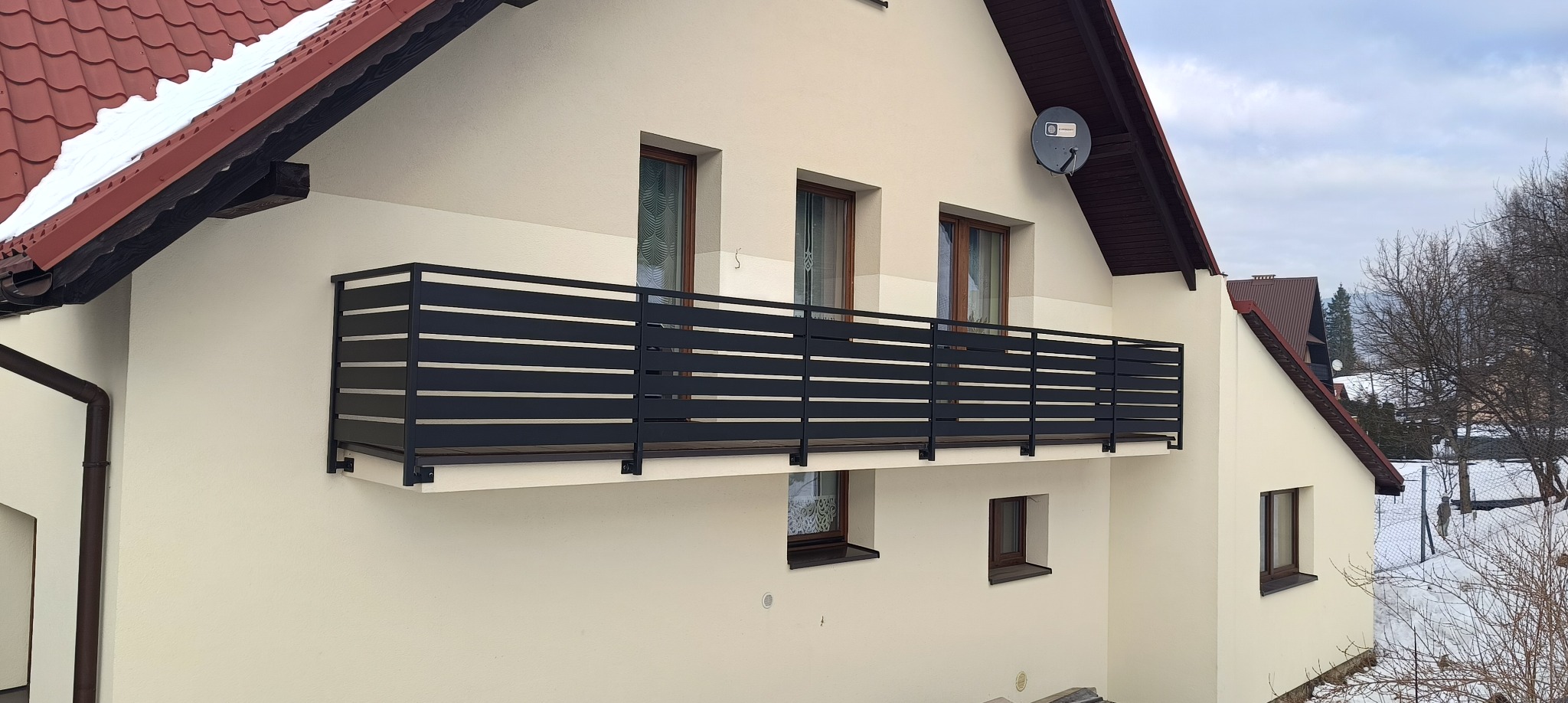 Czarna, pozioma balustrada balkonowa z metalowych profili na tle beżowej elewacji domu z brązowym dachem. Widok z dołu, zima w tle.