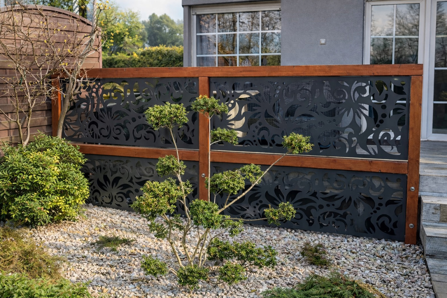 Dekoracyjny panel ogrodowy z wycinanym wzorem kwiatowym, osadzony w drewnianej ramie, otoczony zielenią i białym żwirem. Nowoczesny design.