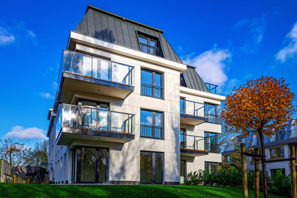 Nowoczesny budynek z balkonami zabezpieczonymi szkłem, na tle błękitnego nieba i zielonego trawnika. Widoczne detale architektoniczne i jesienne drzewo.