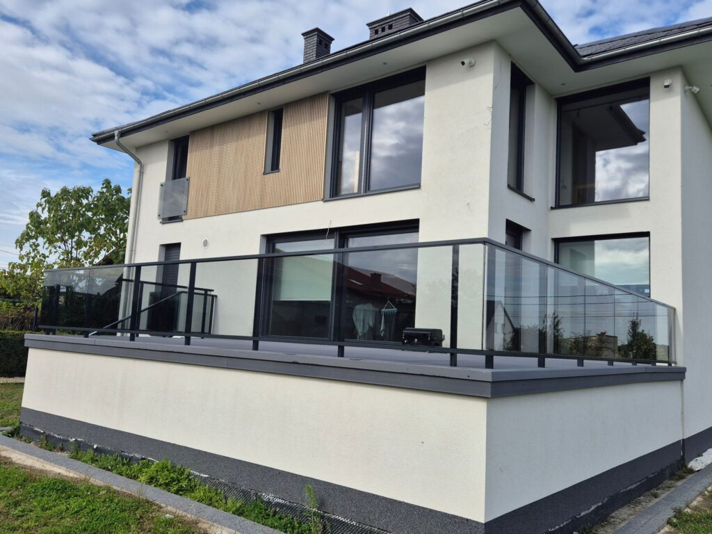 Nowoczesny balkon z czarną, szklaną balustradą w domu z białymi ścianami i drewnianymi akcentami. Widok z dołu, błękitne niebo z chmurami w tle.