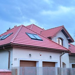 GreenCert Wojciech Mroczkowski - Dom jednorodzinny z czerwonym dachem i dwoma oknami dachowymi, widoczny zza ogrodzenia. Na elewacji numer 37 i lampy nad bramą garażową. Pochmurne niebo w tle.