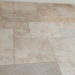 Jarbud - Beżowa podłoga z płytek o zróżnicowanych rozmiarach i fakturach, ułożonych w nieregularny wzór. Widoczne fugi i fragment białej ściany w tle.