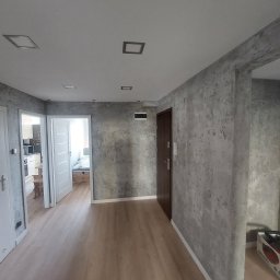 Jarbud - Wnętrze mieszkania po remoncie: szare ściany imitujące beton, drewnopodobna podłoga, białe drzwi i listwy przypodłogowe. Widoczne oświetlenie sufitowe.